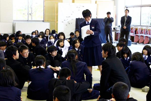 床に座る男女の生徒の中、立っている1人の女子生徒が、紙を片手に話をしている写真