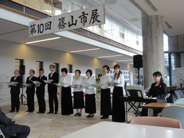 「第10回 篠山市展」の看板の下で黒いスーツに蝶ネクタイの男性と白いブラウスに黒いスカートの女性のコーラス隊が電子ピアノの演奏に合わせて歌を歌っている様子の写真