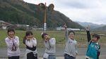 後ろには山々と田んぼが広がっており、その前の道路に西紀 西紀小学校4年生の児童が5名並んでおり女の子3名が笑顔で両手を頬に当てており、男の子2名が右手を上げて人差し指を立ててポースを取っているシーンの写真