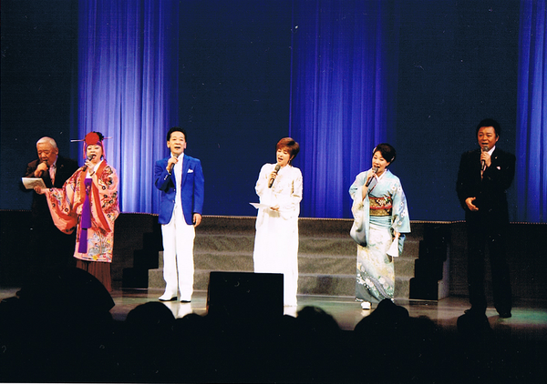 舞台の上で、山口 かおるさん、丹波 ひろしさん、着物を着た女性や、スーツの男性の計6名が、マイクを持ち歌っている写真