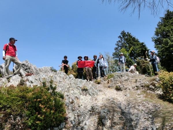ごつごつした岩の上に登山客が休憩しており、その中に赤い布を3人で広げて見下ろしている様子を下から写している写真