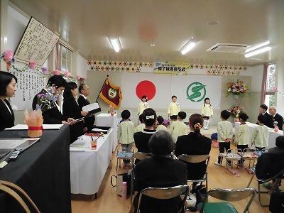 卒園式で卒園生3名が舞台の前に立って在園児と向い合っており、保護者席の後から全体の様子を写している写真