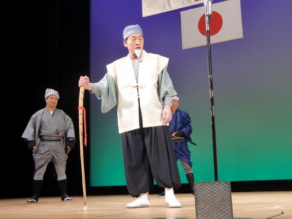 ステージの上で白いひげをつけて、杖を持った男性が水戸黄門の様に2人のお供を従えて、話をしている写真