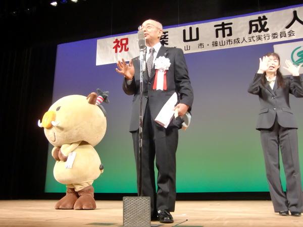 ステージの上で成人式の参加者に話をする市長とその横で手話をする女性と篠山市のいのししのキャラクターが立っている写真