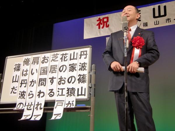 マイクで話をする男性が立っていて、その横に丹波篠山山家の猿が花のお江戸で芝居すると文字の書かれたホワイトボードが置かれている写真