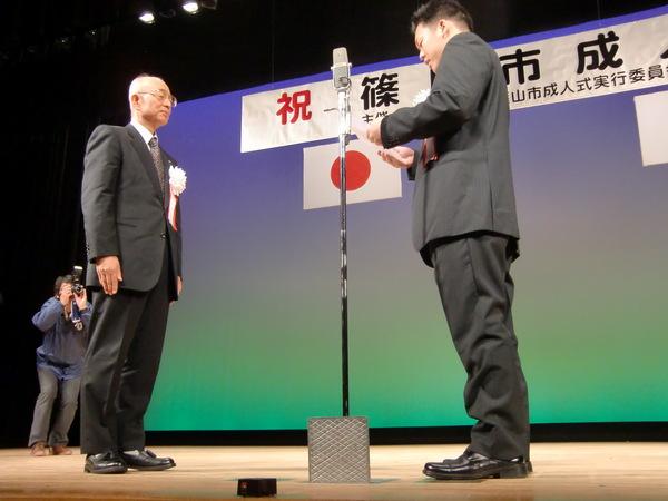 ステージの上で誓いの言葉を述べる新成人とそれを聞いている市長の写真