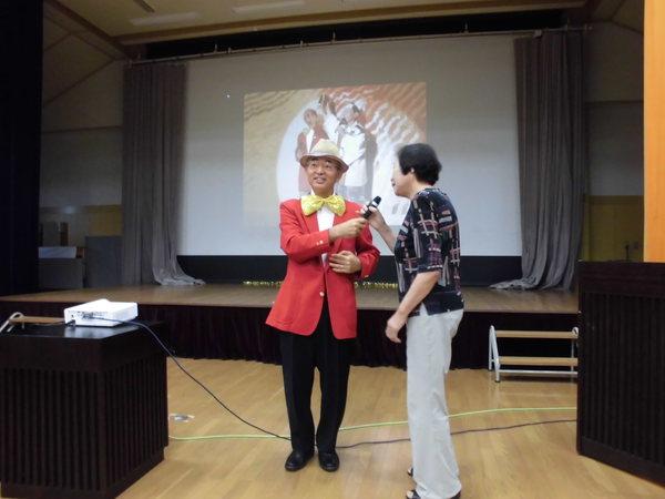 市長が赤いスーツに大きなリボンを胸につけ帽子を被り生徒の方にマイクを向けている写真