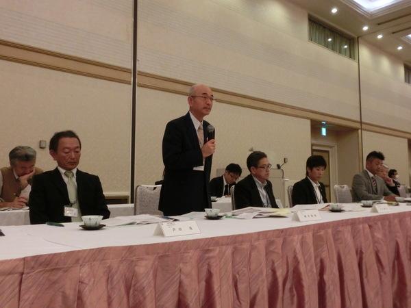 会議にて市長が立ってマイクをもって話をしており、周りの出席者が机の資料に目を通して話を聞いている様子の写真