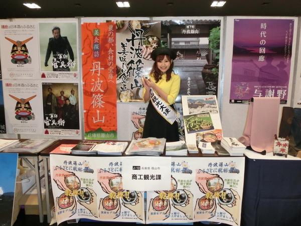 ポスターやパンフレットの前で観光大使がお猪口を持ってPRしている写真