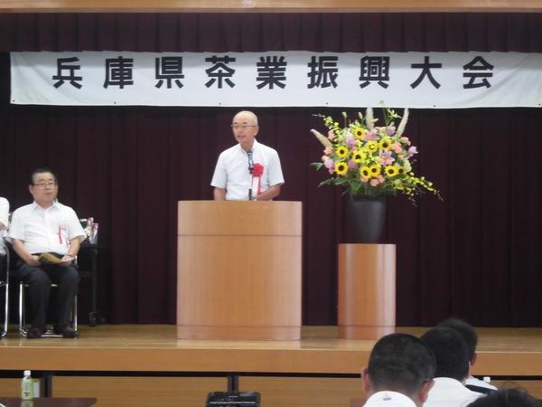 舞台上に「兵庫県茶業進興大会」の横断幕が掛けられており、ひまわりやピンクの壇上花が飾られ、市長が演台の前で話をしている様子の写真