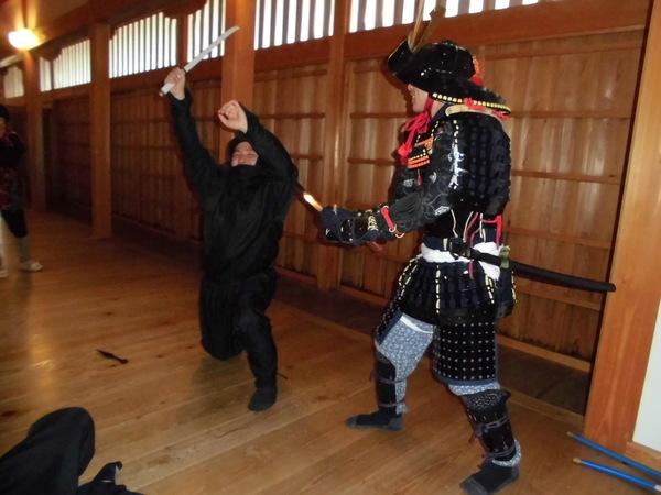 鎧兜姿の男性が、忍者姿の男性を斬る真似をしていて忍者姿の男性が片膝をついている写真