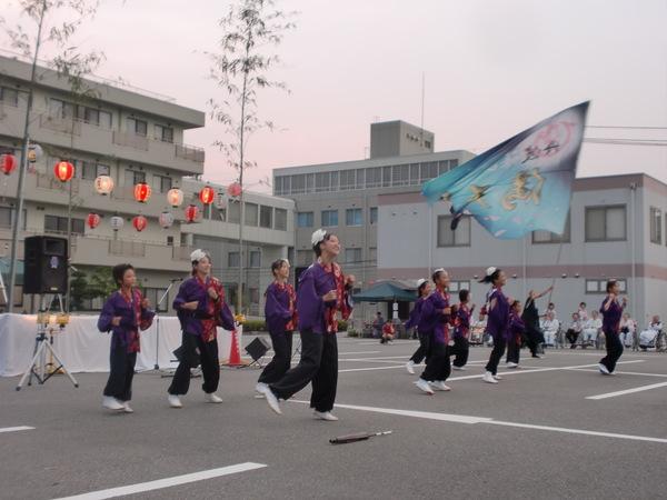 紫の法被を着た女性達が踊り、黒い服を着た人が大きな旗を振っている写真