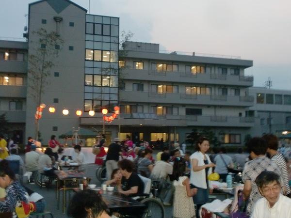 舞台には提灯が灯され、飲食コーナーで沢山の人が座っている写真