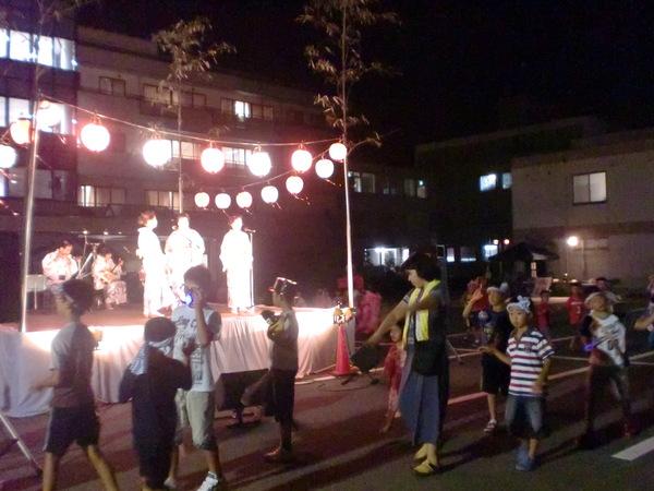 舞台の上で着物を着た人が3人立ってマイクで話し、舞台の下では子供達が踊っている写真