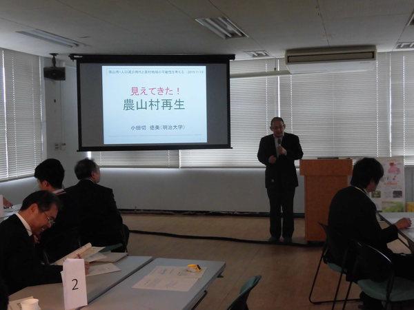 スクリーンには農山村再生とあり、前でマイクで男性が話をし、手前の男性が資料を眺めている写真