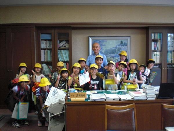 市長室の机の周りに集まる、黄色い帽子をかぶって、ピースサインをする笑顔の小学生の生徒たちと、市長の写真