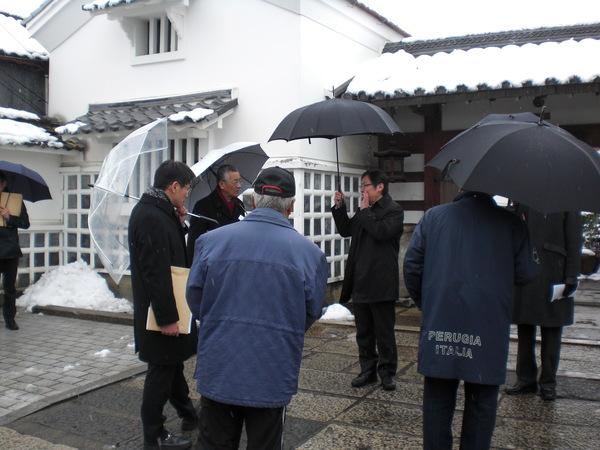 屋根に雪が降り積もる中、全国弁護士市長会に出席された市長の方々が、傘をさして重要伝統的建造物群保存地区の河原町の視察に訪れている様子の写真