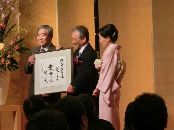 舞台で男性2人が大きな額を持ちその隣で着物を着た女性が立っている写真