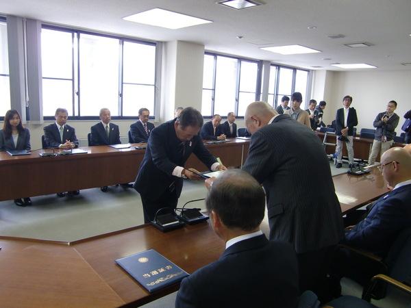 当選された議員さんが座っていて、1名の男性が当選者に当選証書を渡している写真
