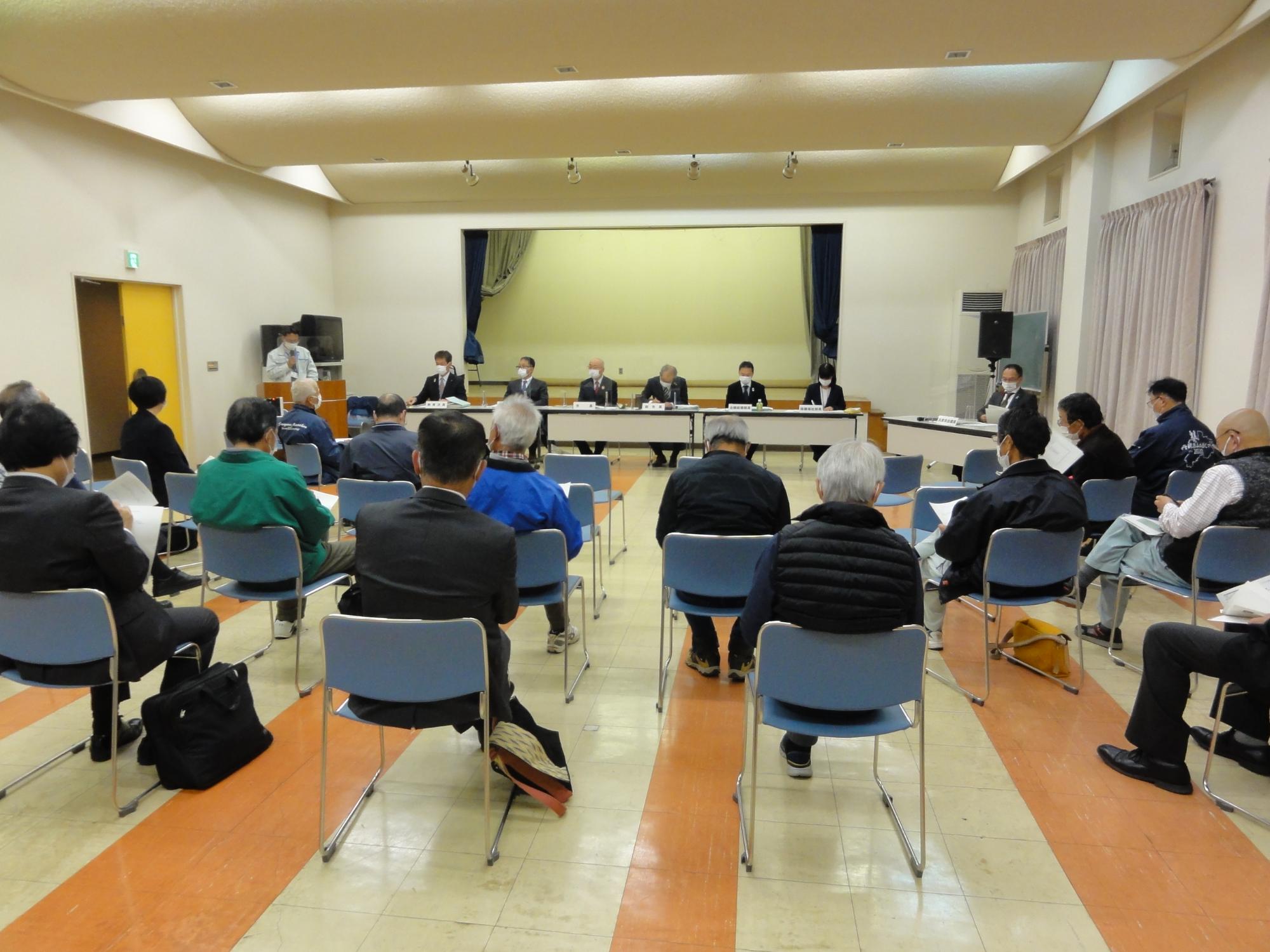 会議室の前方に、市幹部と県会議員が並んで座っている。後方に椅子が3列ほど並んでいて出席者が資料を持って座っている。