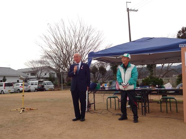 紺色のスーツ姿の市長がマイクを手に持ち挨拶をしている隣で白地にエメラルドグリーン色の線が入った帽子を被った男性が笑顔で立って話を聞いている写真