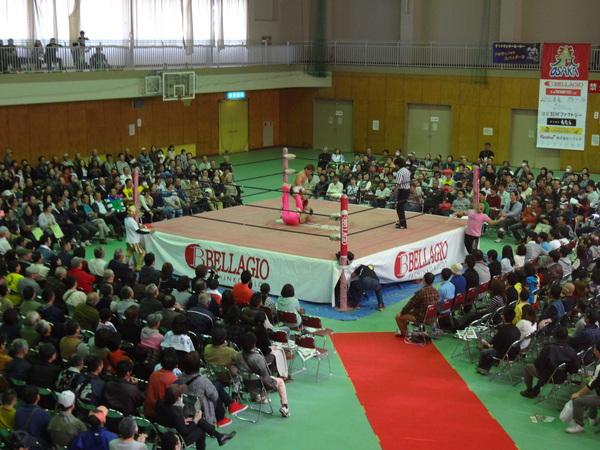 リング場で、ピンクのズボンを履いた男性と黒いパンツを履いた男性が対戦しており、リング場の周りに作られた観客席には満員のお客さんが座って試合を見ている様子の写真