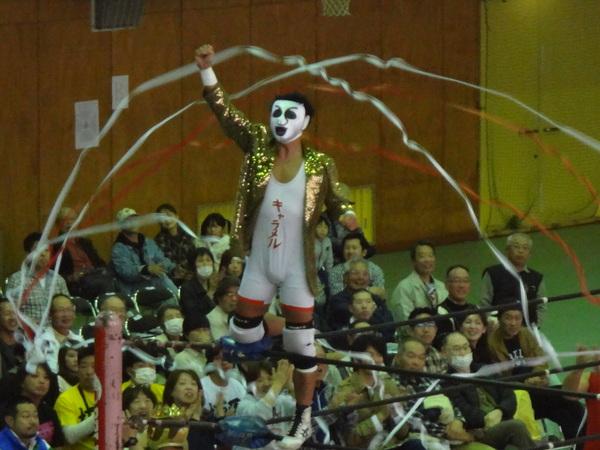 「キャラメル」と書かれた衣装を着て、白いマスクをかぶった男性が、リンクのロープに足をかけてガッツポーズをして立っており、観客席からリボンテープが飛び、拍手をされている様子の写真