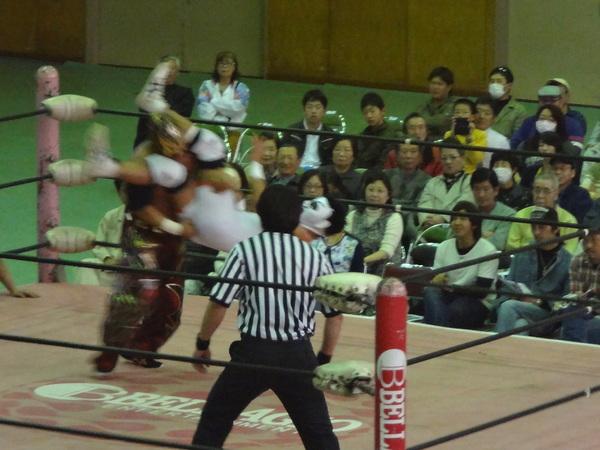 リング場で、白いマスクを付けた男性が、技をかけられ投げ飛ばされている瞬間の写真