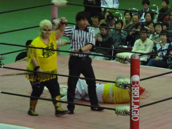 リンクの上で黄色い極悪と書かれたティーシャツを着たダンプ松本さん右手を上げ、審判が勝利のジャッジをしており、対戦相手がリンクに倒れている様子の写真
