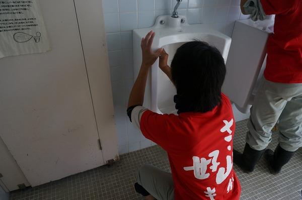 「ささ山が好き」のバックプリントされた赤いTシャツを着た男性が男子トレイの便器を磨いている様子の写真
