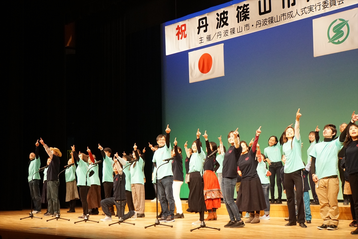 田園交響ホールの舞台で、水色のTシャツや黒のTシャツを着た人たちが左の人差し指を高く掲げ、指さしている