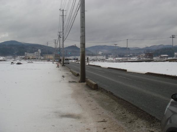 広場には雪が積もり、道路を車が走り、遠くに青信号と山が見えている写真