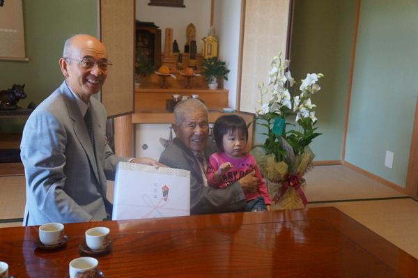 大西さんがひ孫さんを膝の上に抱いて、市長が記念品を渡す様子の写真