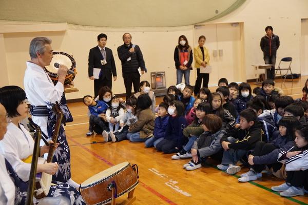 浴衣を着た男性が締太鼓を手に持ちマイクで話をしており、それを興味深く座って聞いている子供たちの写真
