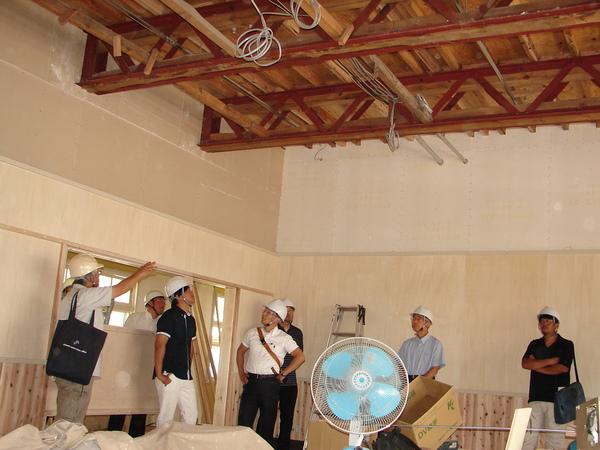 扇風機が置かれた、工事中の一室で、男性が指さす方を見ている、市長と関係者の写真