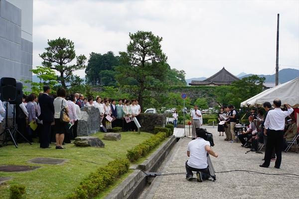 曇り空の下、芝生で合唱するグループをテレビカメラで撮影をしている写真
