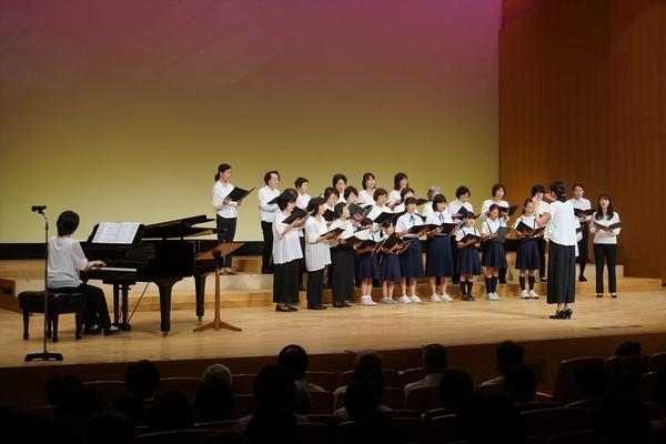 舞台の上で制服を着た合唱団が、楽譜を持ってピアノ伴奏に合わせて歌っている写真