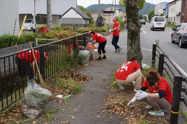 お揃いの赤いシャツを着た男女が歩道に落ちている落ち葉やごみを拾っている写真