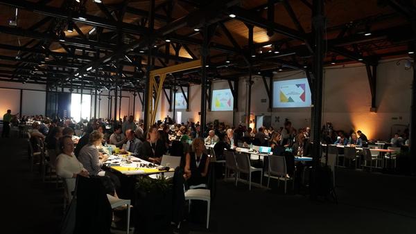 室内が暗い会場で白いテーブルが置かれていて、大勢の人達が座っている写真