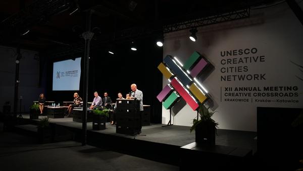 壁や床が黒色の会場の舞台の演台に男性が立って話をしている写真