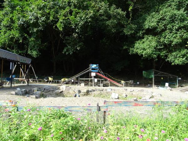 木々の下に手作りの木で作られてた滑り台が写っており、中央には石がが置かれ大きな穴のような物が空いており、手前にはピンクの草花が咲いている写真