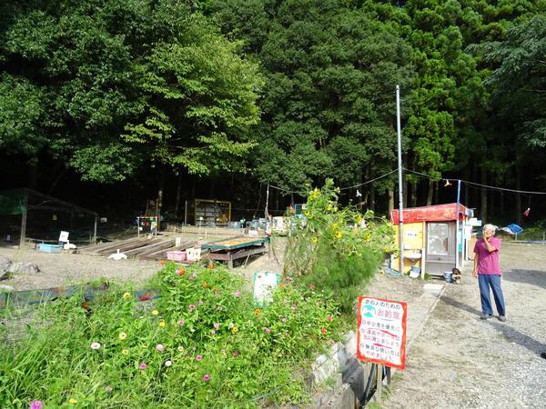 奥は森になっており、手前の広場に手作りの遊具が置かれており、赤い屋根の小屋や手前にはお約束の書かれている看板が表示されており、紫色の洋服を着た男性が立って写っている写真