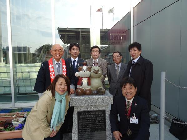 犬山市役所前のキャラクターわん丸くんとまるいのが飾られた前で市長やスーツ姿の男性と女性で笑顔で記念写真