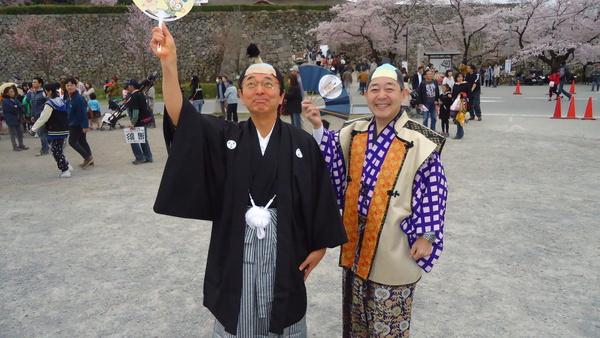 祭り会場で紋付袴を着てちょんまげのカツラを被った市長が右手に団扇を持ち議長は青色の扇子を片手に上げた写真