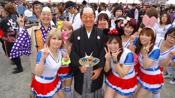 紋付袴を着てちょんまげのカツラを被った市長と議長と白地に青地の袖赤いフリルの衣装を身に着けた女性達に囲まれて笑顔で記念撮影した写真