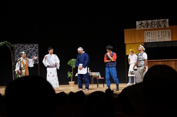 舞台にセットされた食堂に殿様、襷をかけた浴衣姿の男性、ねじり鉢巻きに前掛け姿の店のご主人、とび職人らが立って話をしている写真