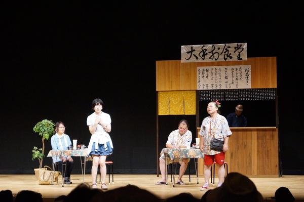 舞台にセットされた食堂でねじり鉢巻きをしているお店の主人とお客が2組がテーブルに居る写真