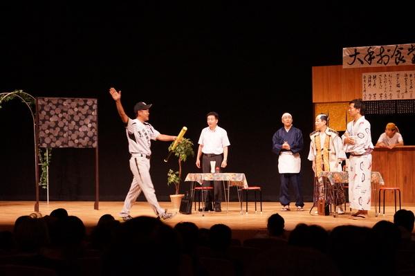 舞台にセットされた食堂で野球のユニホーム姿で左手にバットを持ち右手を上げている男性と殿様が話をしているのをねじり鉢巻きに前掛け姿の店のご主人らが見ている写真