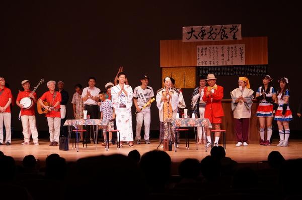 舞台にセットされた食堂で劇に出演した市長らが楽器の演奏に合わせて手拍子している写真