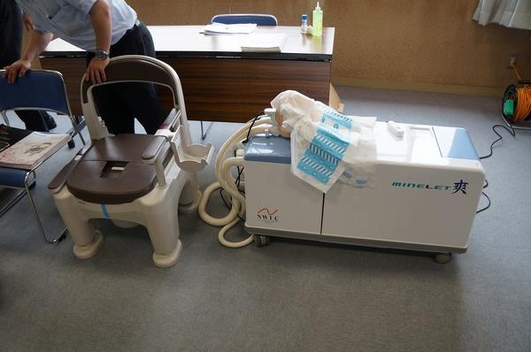 ポータブルトイレの便器の横に置かれている自動排泄物処理装置ロボット「マインレット爽」の写真